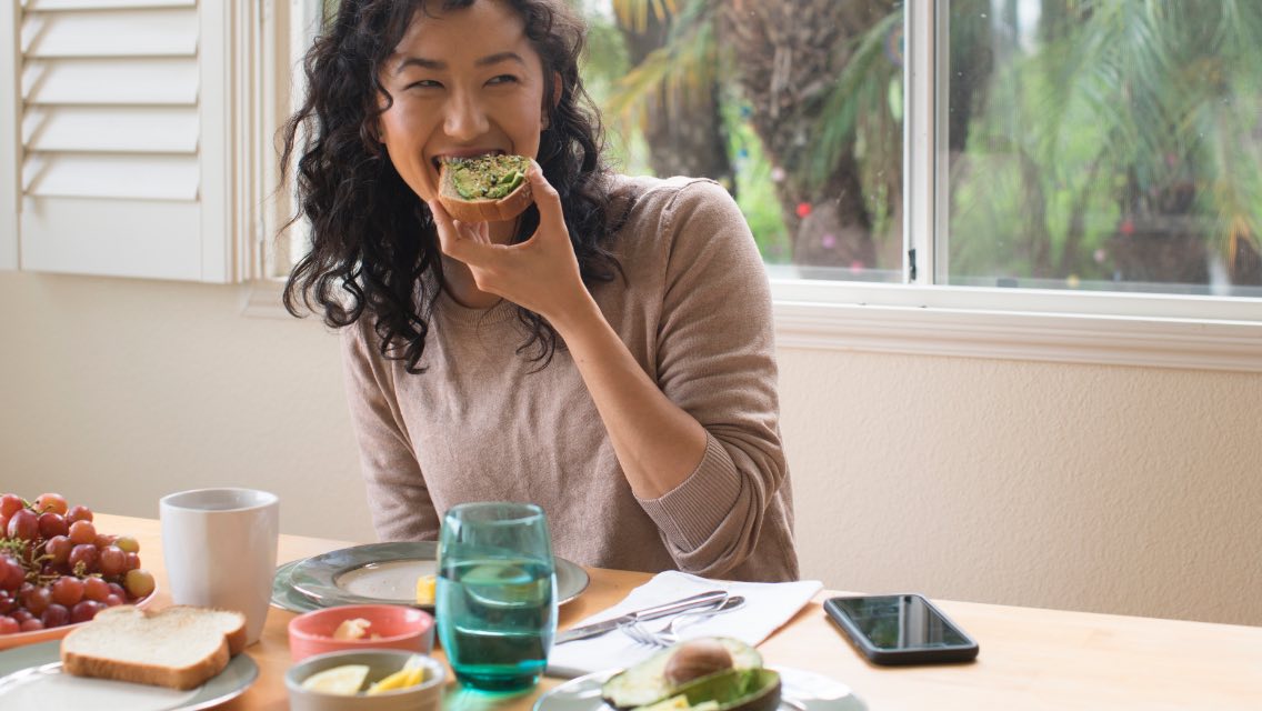 A woman smiling while eating avocado toast in the kitchen.