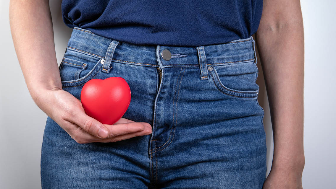 A woman holds red wooden heart next to her pelvis.