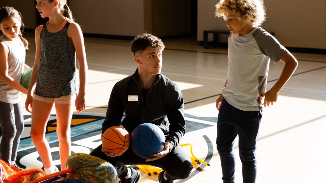 A Life Time team member holding sports equipment, including a basketball, while talking to younger kids in a gym.