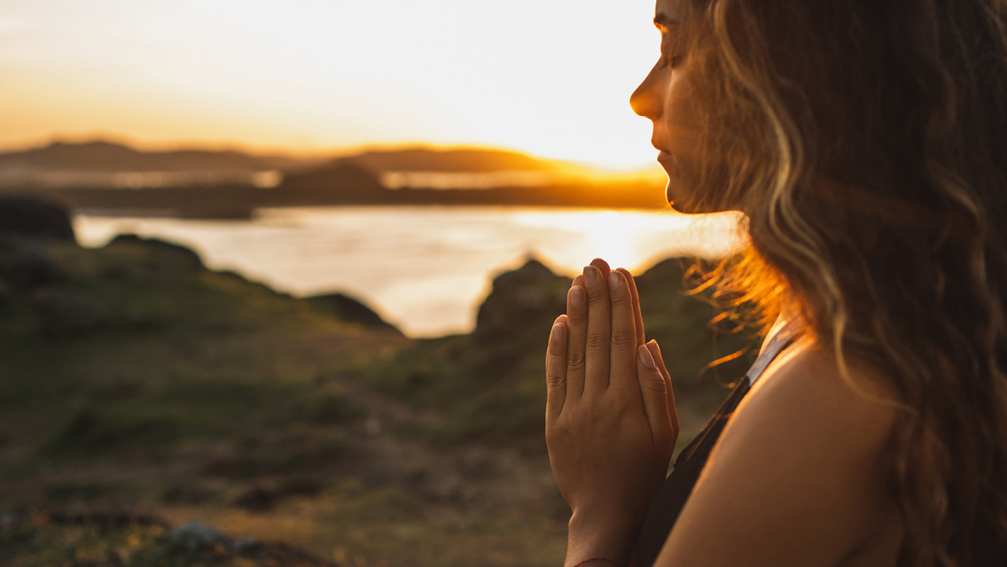 a woman holds hands in prayer position with the sunsetting behind her