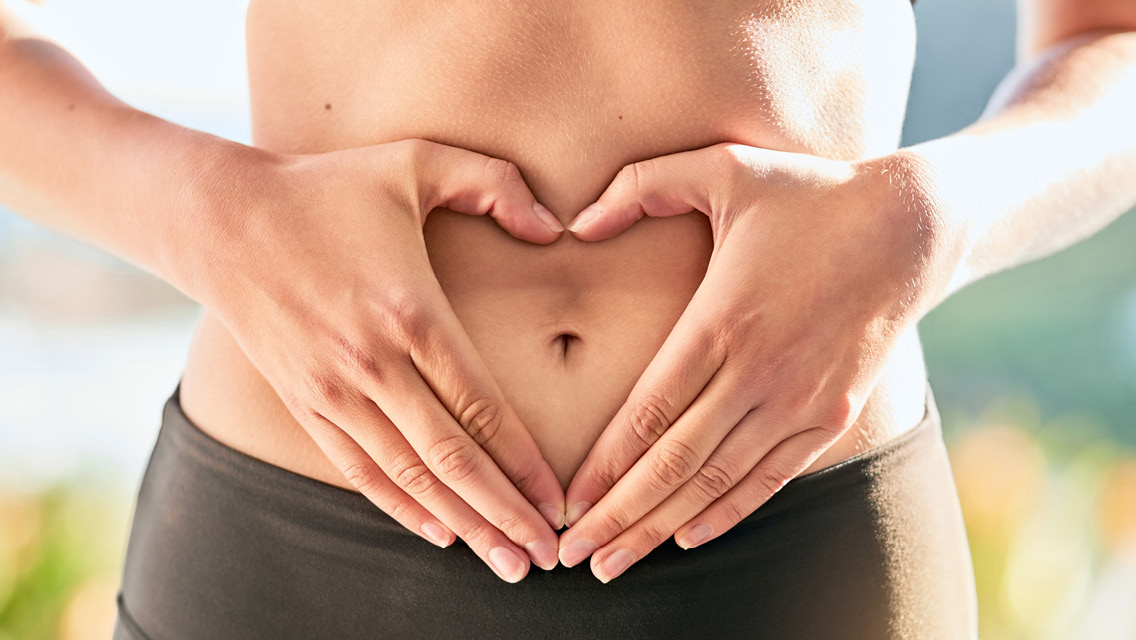 a woman holds her hands in a heart shape around her belly button