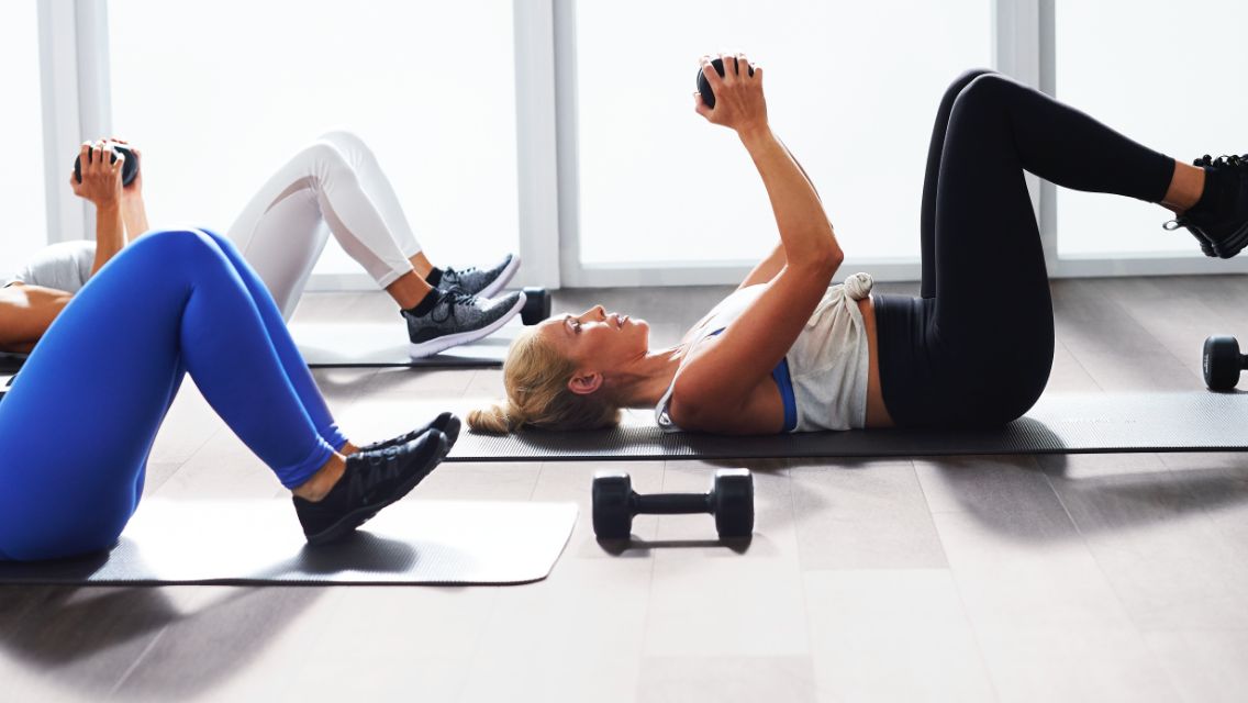 Three women working out together in a health club, lying on their backs using dumbbells.