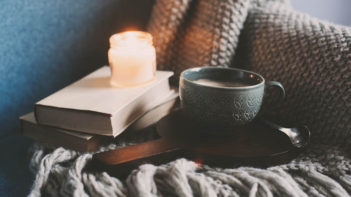 a candle, book and cup of warm cocoa rest on a blanket