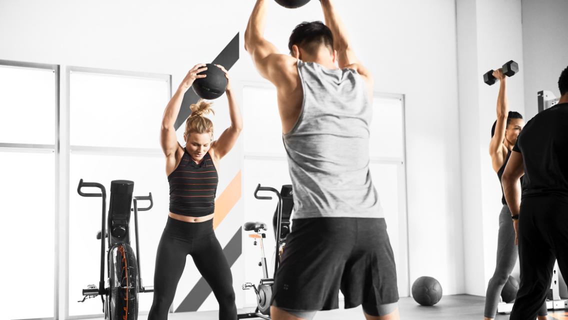 Individuals working out in a group training class setting, using medicine balls and dumbbells.