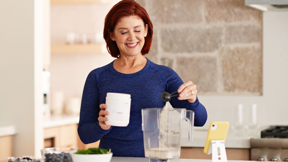 A woman in her kitchen scooping supplement powder into a blender.