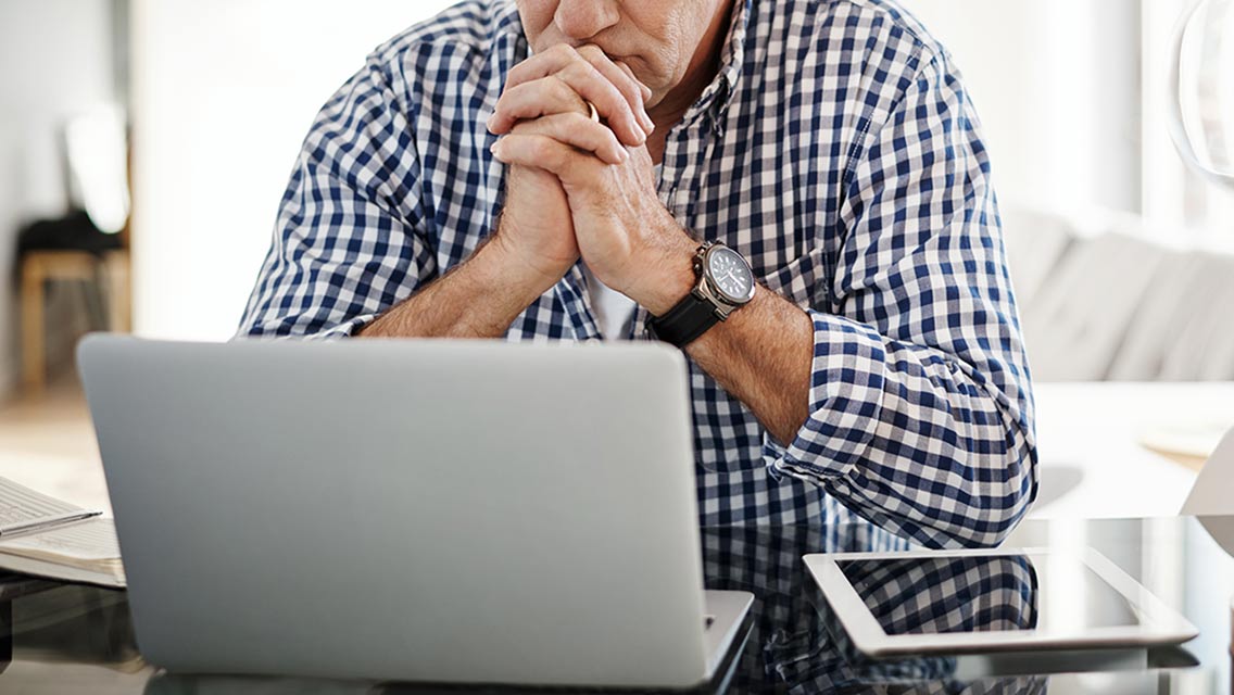 a senior man sits concernedly in front of his computer with hands folded