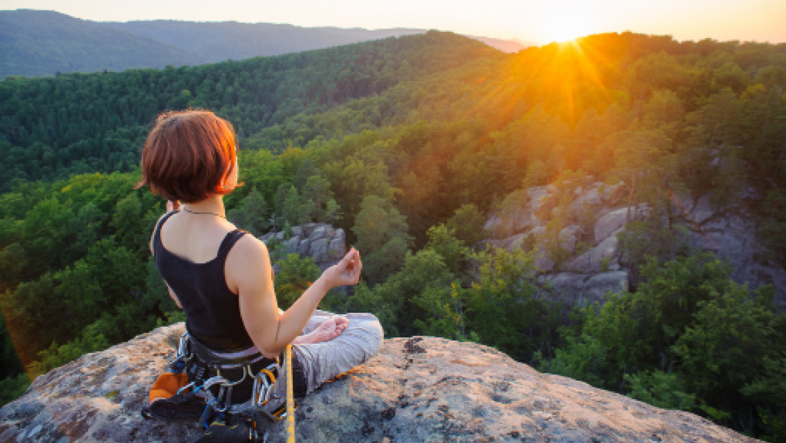 a woman meditates on top of a cliff with her climbing appartus attached