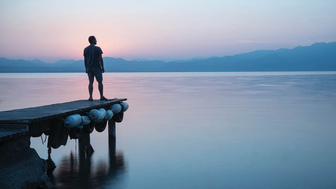 a silhouette of a man standing at the end of a dock with a pastel sunrise