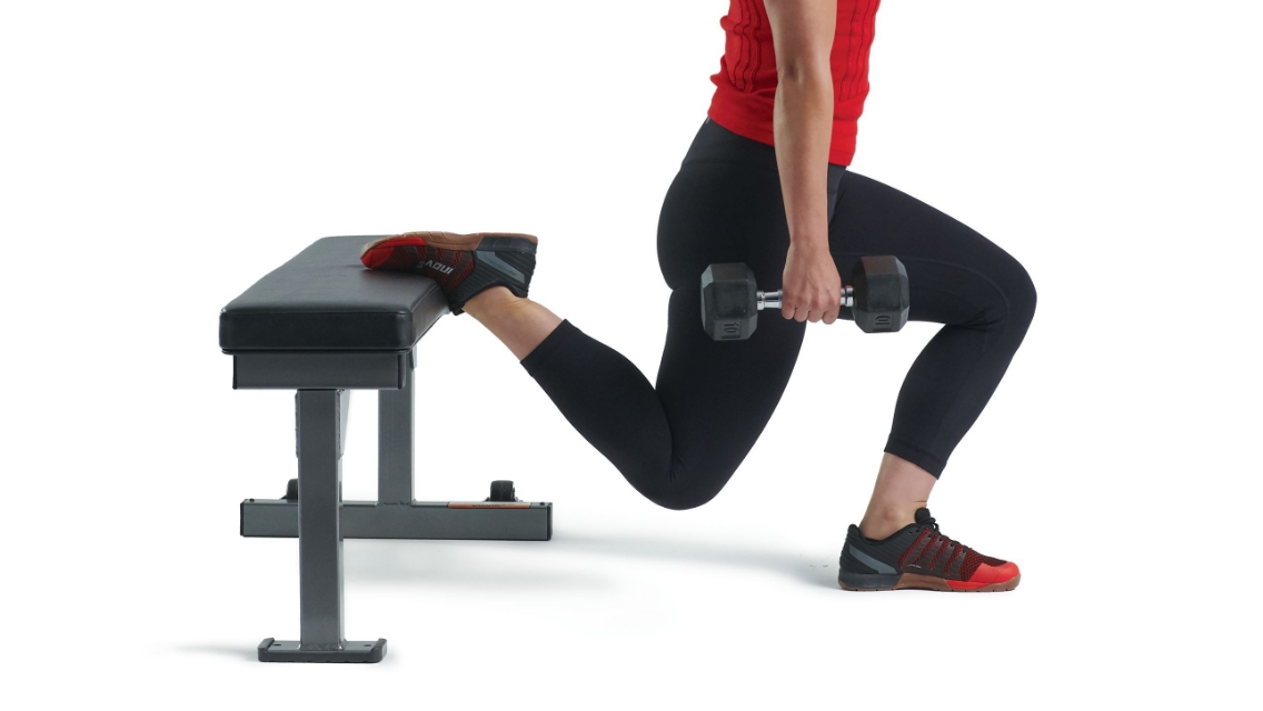 A woman doing an elevated squat with one leg on a bench and one on the floor while holding a dumbbell.