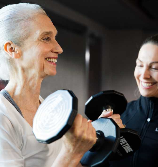 A Life Time trainer working with an older adult woman who is lifting dumbbells.