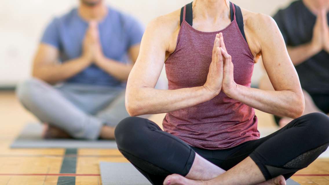 Three people in a yoga class sitting cross-legged with arms at heart center.
