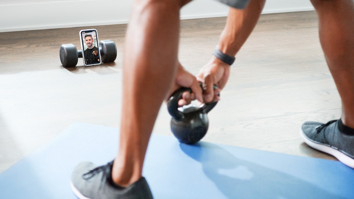 A person doing a workout with a kettlebell at home while communicating virtually on their phone with a trainer.