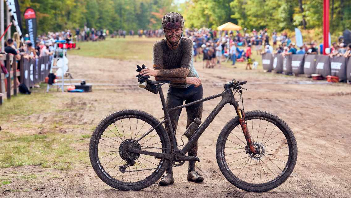 Alexey Vermeulen standing next to his bike at a finish line covered in mud.