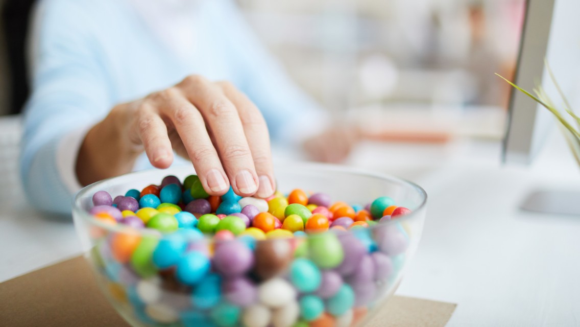 A person sticking their hand into a bowl of candy while working at a desk.