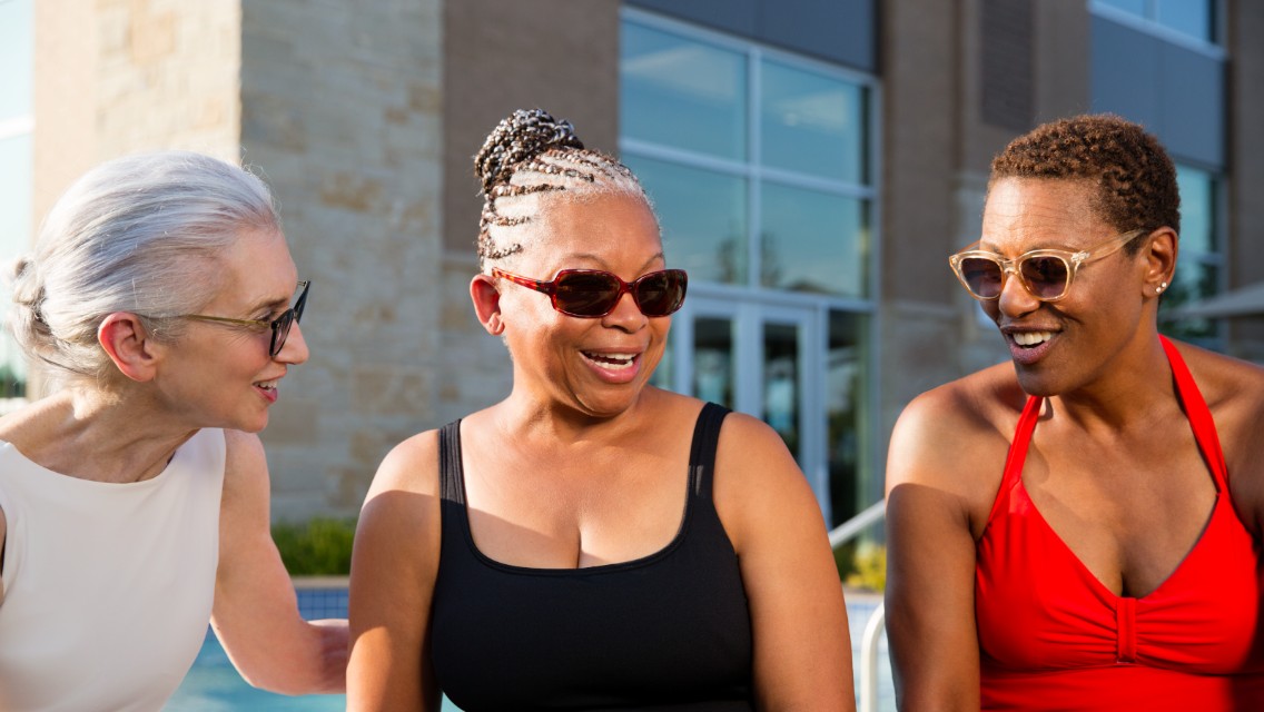 Three older adult women smiling and talking outside by the pool.