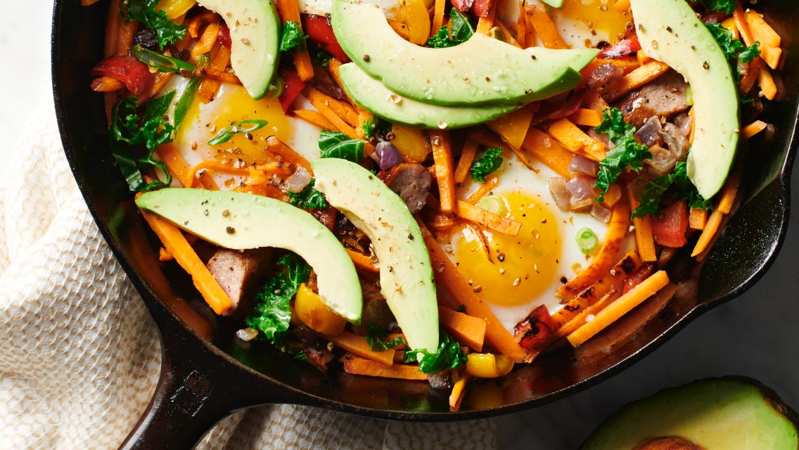 a cast iron skillet with eggs, sliced avocados, kale, meat and sweet potatoes