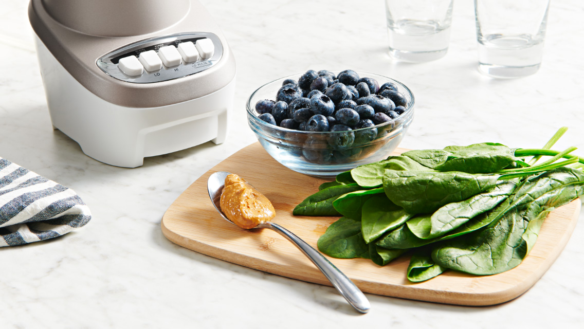Blender, nut butter, blueberries and spinach laid out on a counter.