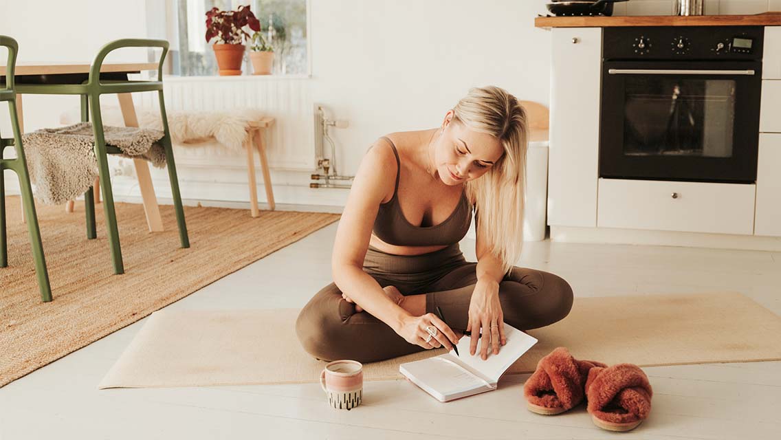a woman in athletic apparel sits on her yoga mat in her home writing in a fitness journal