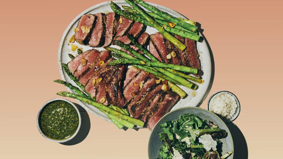 Garlic-seared steak with asparagus on a plate next to a bowl with a side salad and smaller bowls of pesto and parmesan.