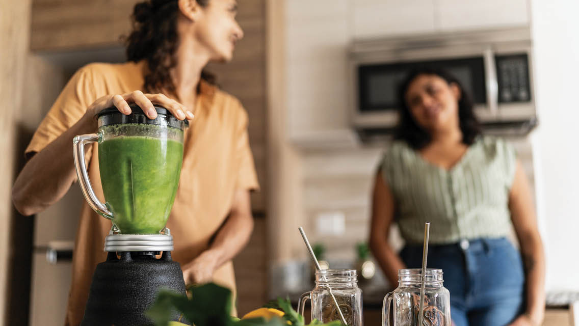 two women with different body sizes are in a kitchen making a green smoothie
