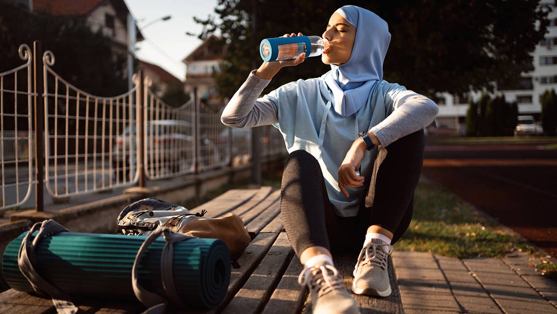 a woman wearing a Hijab sits outside drinking water