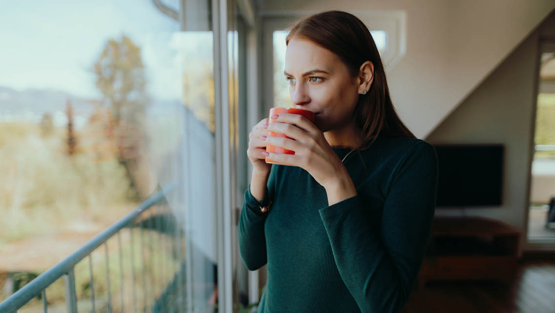 a woman stands drinking a cup of coffee while looking out the window
