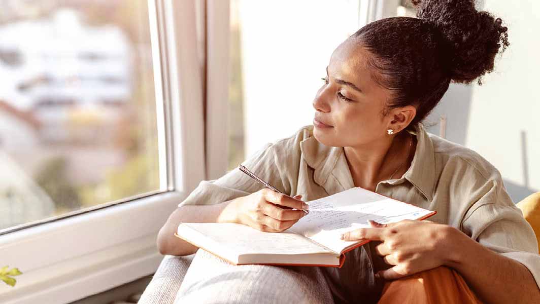 a woman looks out the window while writing in a journal.