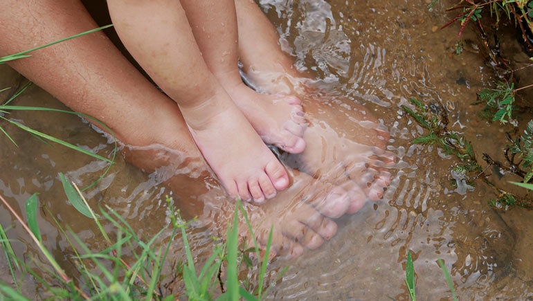 a set of adult feet and toddler feet sit together in a stream