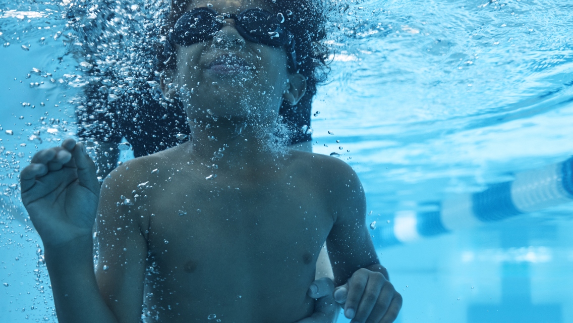 A young bowl swimming under the water in a pool with a swim instructor by his side.