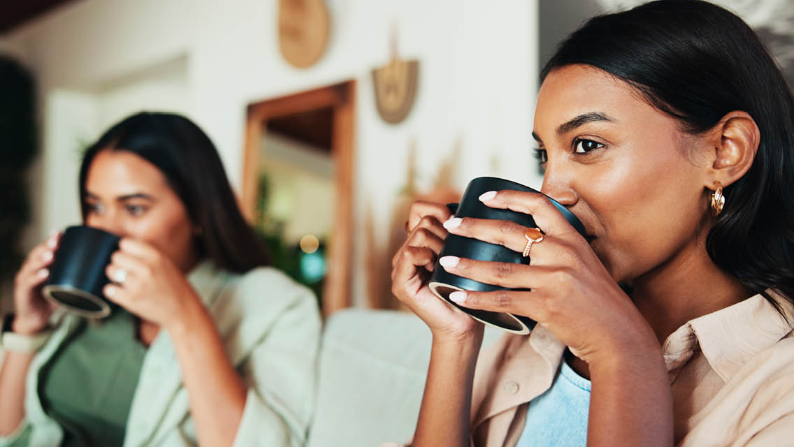 two women sit on a couch drinking coffee staring straight ahead