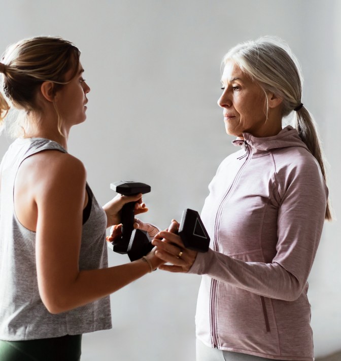 An active older adult woman holding dumbbells while talking to a trainer.