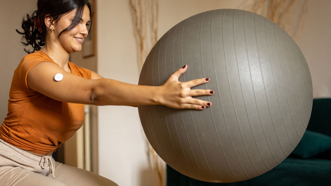a woman holds an exercise ball while wearing a glucose monitor