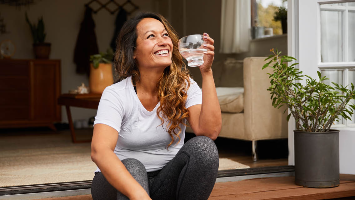 a woman smiles while sitting outside drinking a glass of water