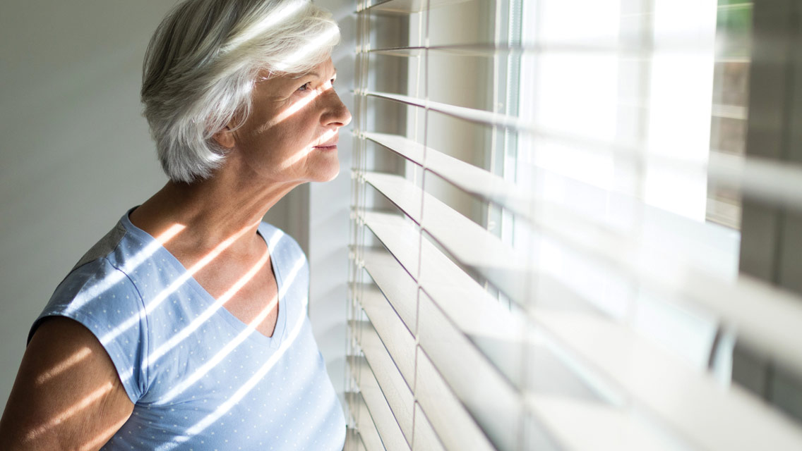 An older woman looks through the blinds out the window.