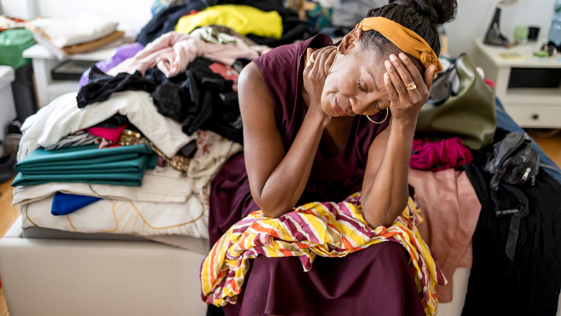 a woman sits looking emotionall upset with a huge stack of clutter on her bed