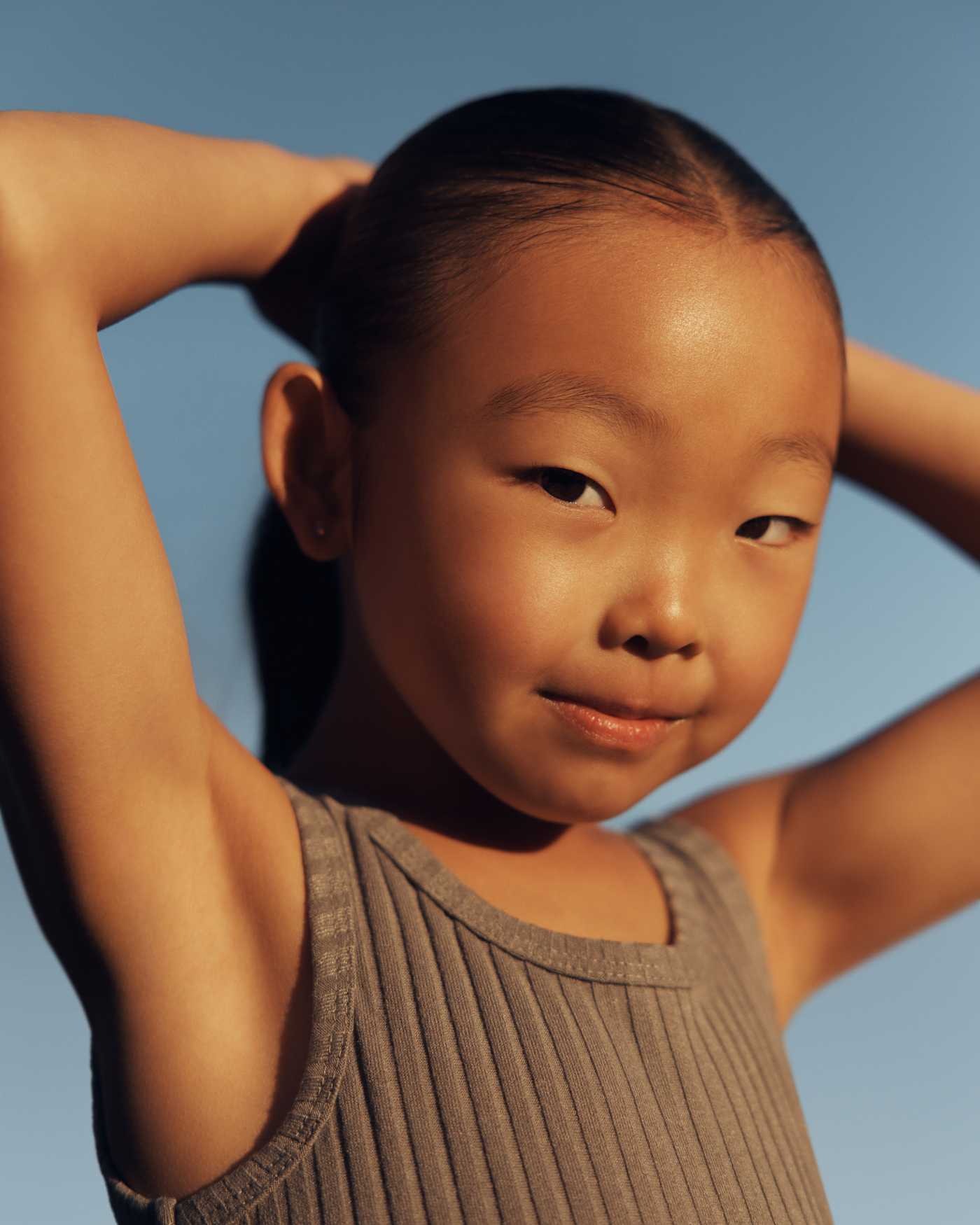 A young girl with her hands behind her head outside.