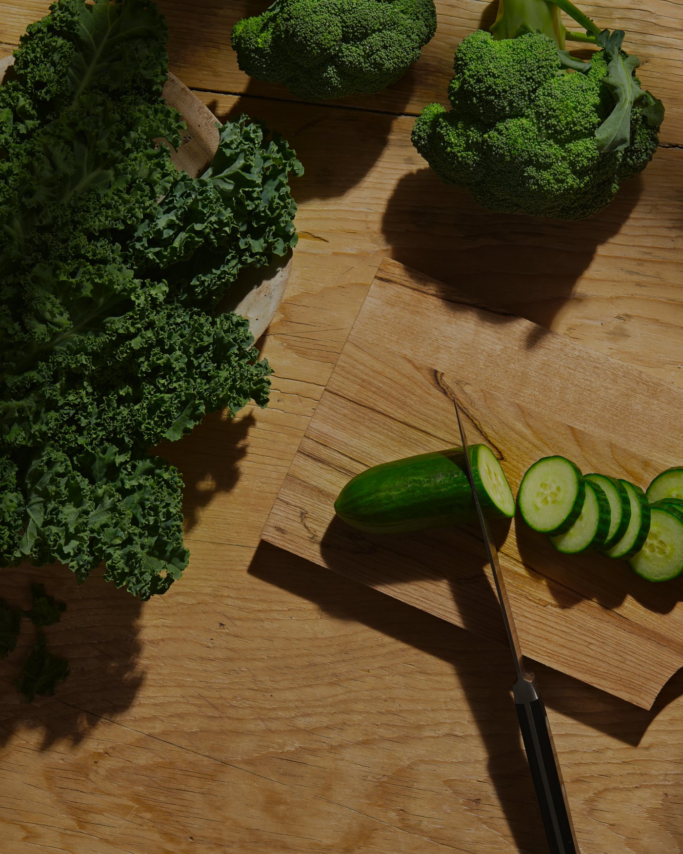 A cutting board with a cucumber being chopped, alongside kale and broccoli.