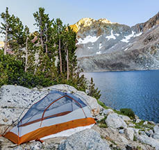 Beautiful campsite on rocky ledge next to alpine snow melt lake. Last rays of sunlight glance mountain tops in the background