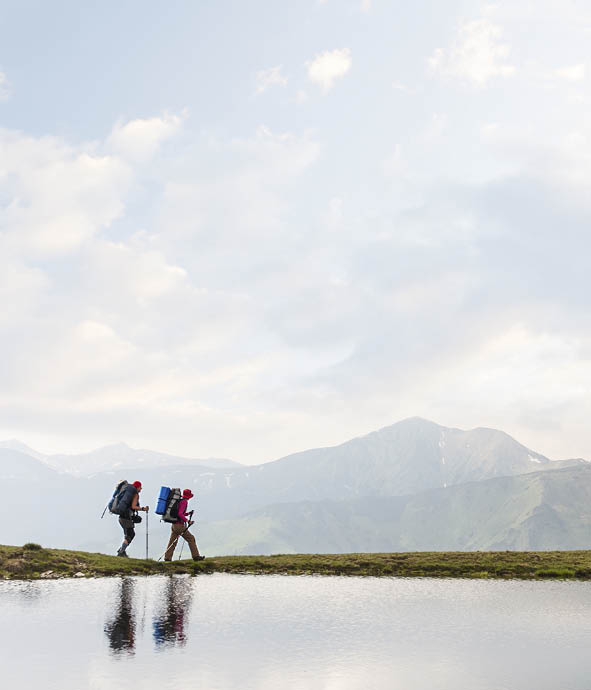 Hikers passing by a clear calm lake reflecting the sky in the mountains
