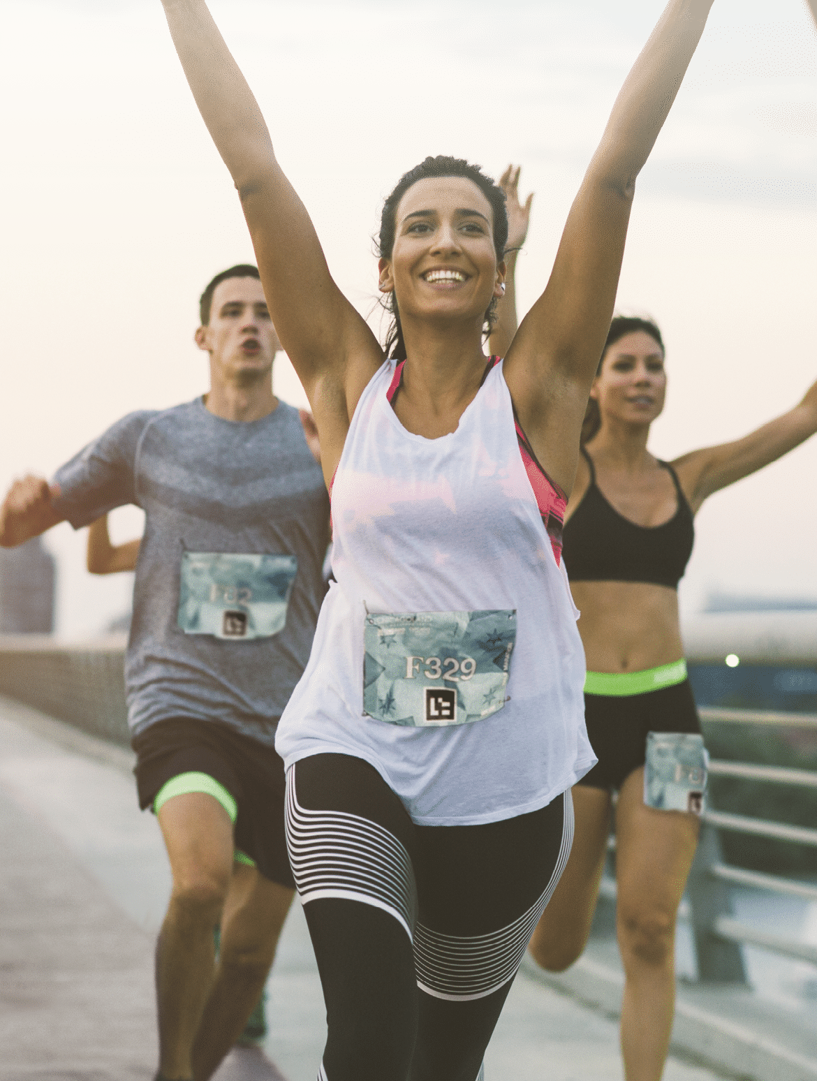 Runners running marathon in the city  They are running with arms raised over the bridge at sunset  Selective focus on beautiful brunette woman running and smiling  Wearing numbers on their sport clothes   Running through the finish with arms raised 