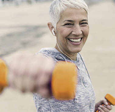 Portrait of cheerful aged woman Fighting breast cancer in fitness wear exercising with pink dumbbells in the city during cloudy day  Front view of Happy Gray hair female doing dumbbell curls in the fresh air  Smiling, Joyful senior, short hair,  lady exercising with weights outdoors  Health and sport 