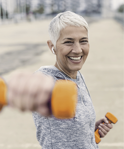 Portrait of cheerful aged woman Fighting breast cancer in fitness wear exercising with pink dumbbells in the city during cloudy day  Front view of Happy Gray hair female doing dumbbell curls in the fresh air  Smiling, Joyful senior, short hair,  lady exercising with weights outdoors  Health and sport 