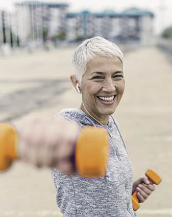 Portrait of cheerful aged woman Fighting breast cancer in fitness wear exercising with pink dumbbells in the city during cloudy day  Front view of Happy Gray hair female doing dumbbell curls in the fresh air  Smiling, Joyful senior, short hair,  lady exercising with weights outdoors  Health and sport 