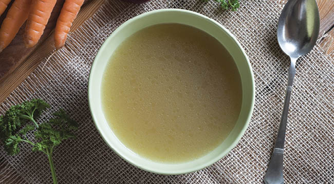 Chicken stock in a green soup bowl, with carrots, onions and parsley in the background, top view