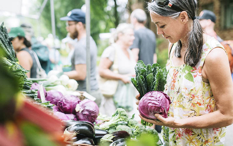 A woman in her 50 s shops in a local outdoor agriculture market with fresh  organic local fruits and vegetables  She smiles as she compares different vegetables for ripeness  Horizontal image with copy space 