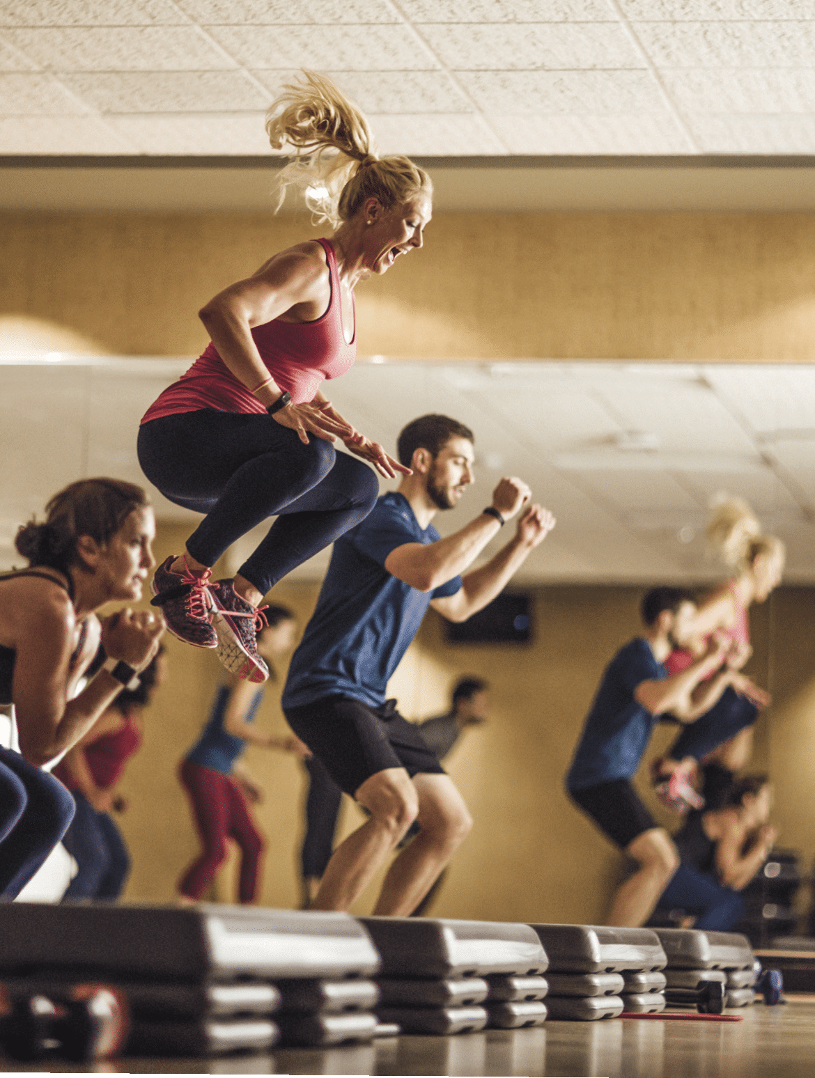 A group of people doing squat jumps in front of exercise steps in a dimly lit group fitness studio