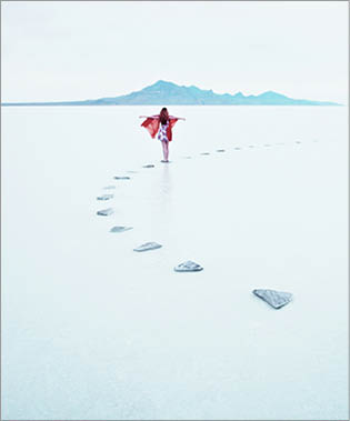 Woman standing on stone pathway in lake with arms outstretched and scarf blowing in wind