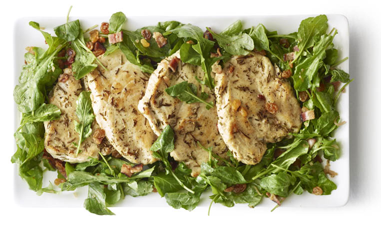 Overhead of rosemary chicken paillards seasoned with rosemary and shallots over lettuce  bacon and golden raisins on a white rectangular plate on a white surface 