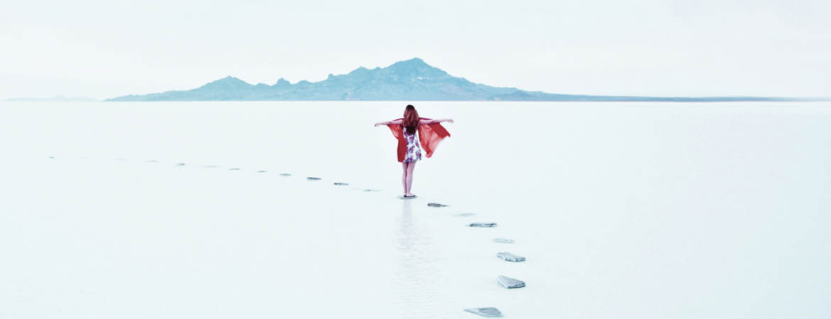 Woman standing on stone pathway in lake with arms outstretched and scarf blowing in wind
