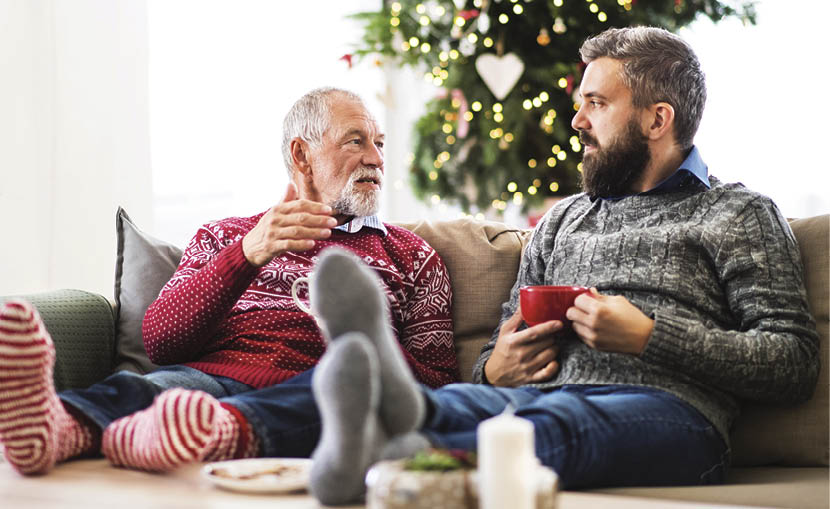 A senior father and adult son with feet on a coffee table sitting on a sofa at home at Christmas time  talking 